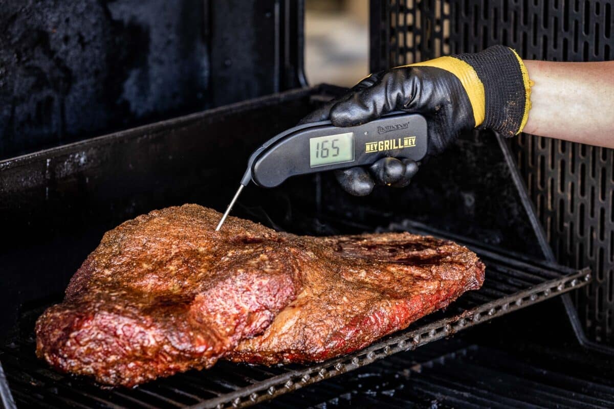 A hand holding a thermometer checking the temperature of beef on a smoker. The temperature on the screen reads 165 degrees F.