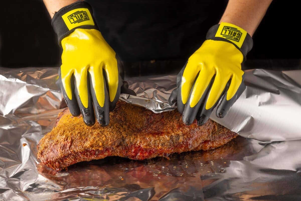 Gloved hands wrapping a brisket in aluminum foil.
