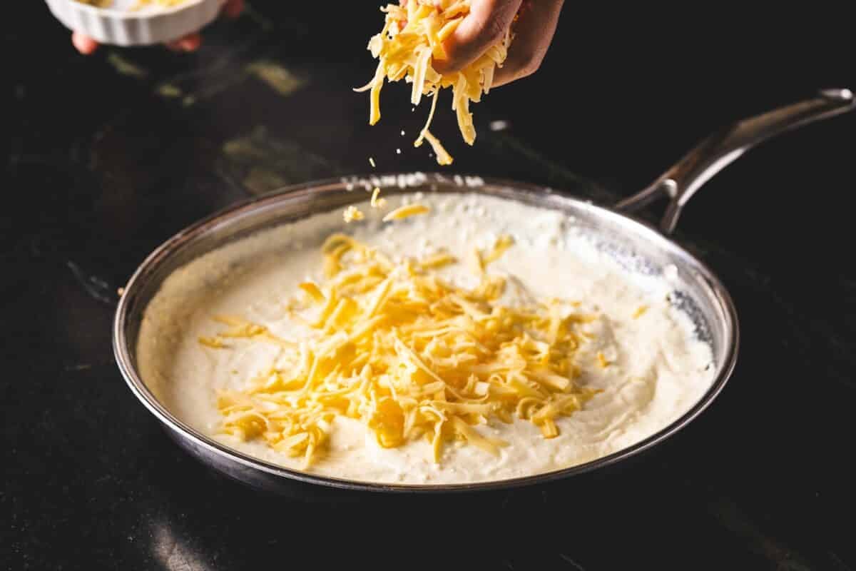 A hand sprinkling shredded cheese into a sauce in a pan below.