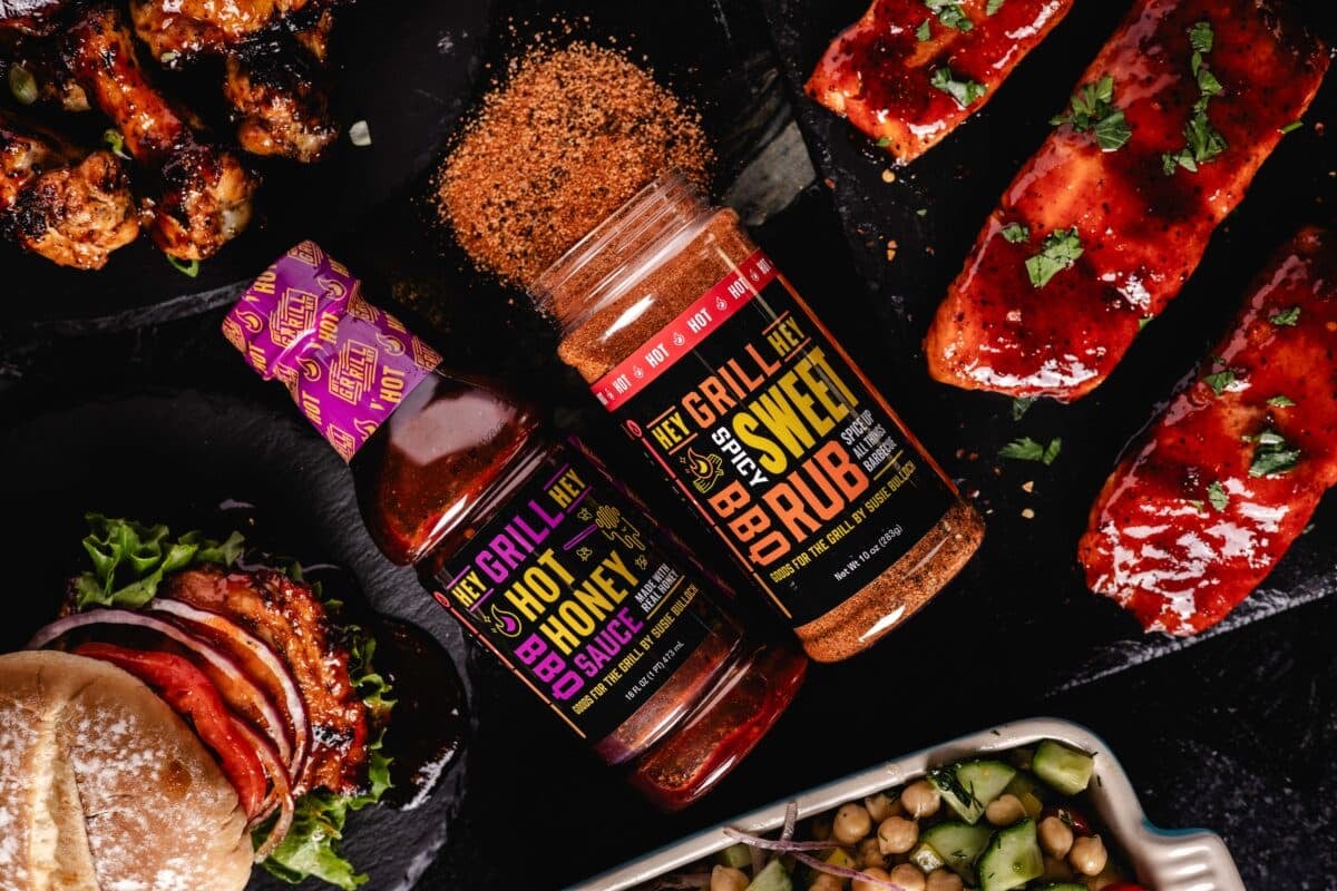 Bottles of Spicy Sweet Rub and Honey BBQ Sauce on a table surrounded by grilled meats.