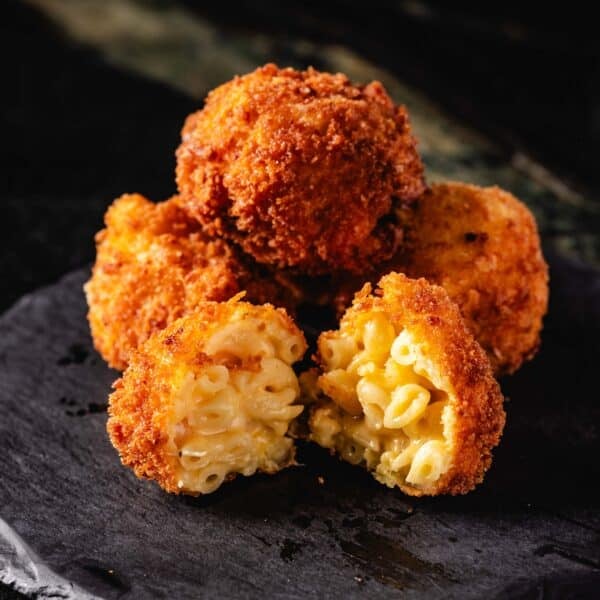 A black platter piled with mac and cheese bites. One is broken open, revealing the creamy mac inside.