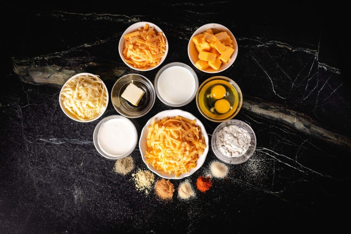 An overhead shot of piles of individual seasonings next to bowls of shredded cheeses and liquid ingredients on a countertop.