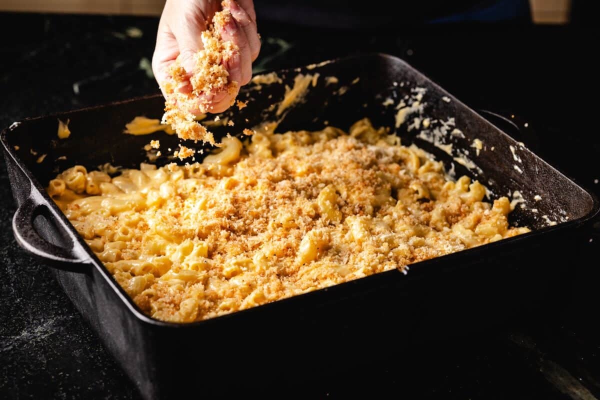 A hand sprinkling breadcrumbs onto a mac and cheese casserole in a cast-iron pan before it gets smoked.