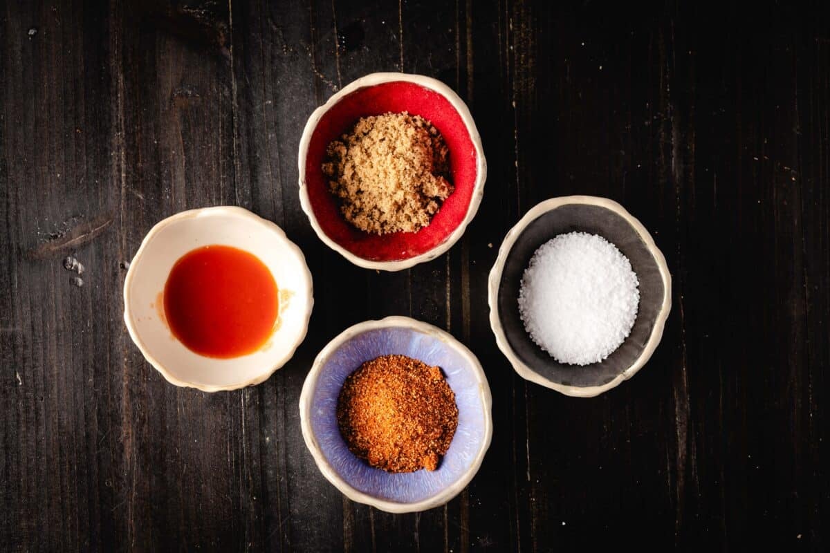 An overhead shot of four bowls holding salt, hot sauce, a BBQ rub, and brown sugar.
