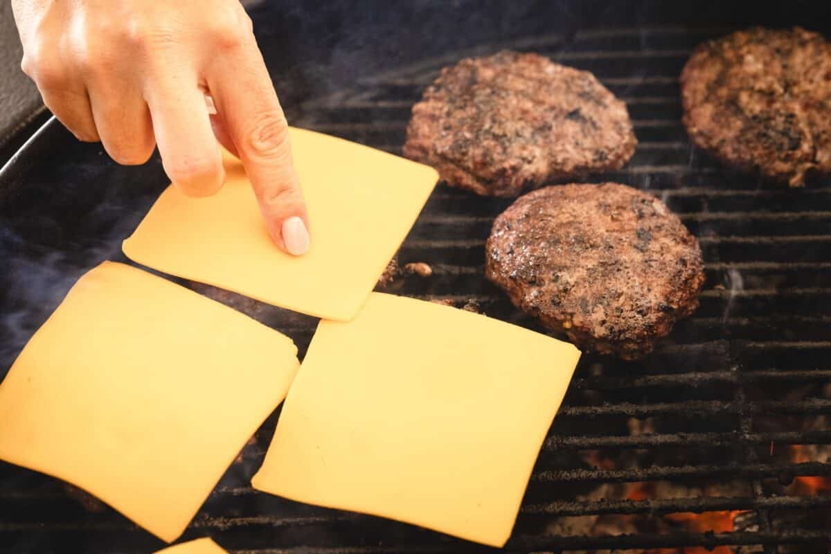 A chef's hand adding cheese slices to burger patties cooking on the grill.