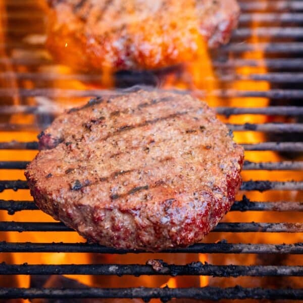 A closeup shot of two burgers being grilled over charcoal with flames rising around them.
