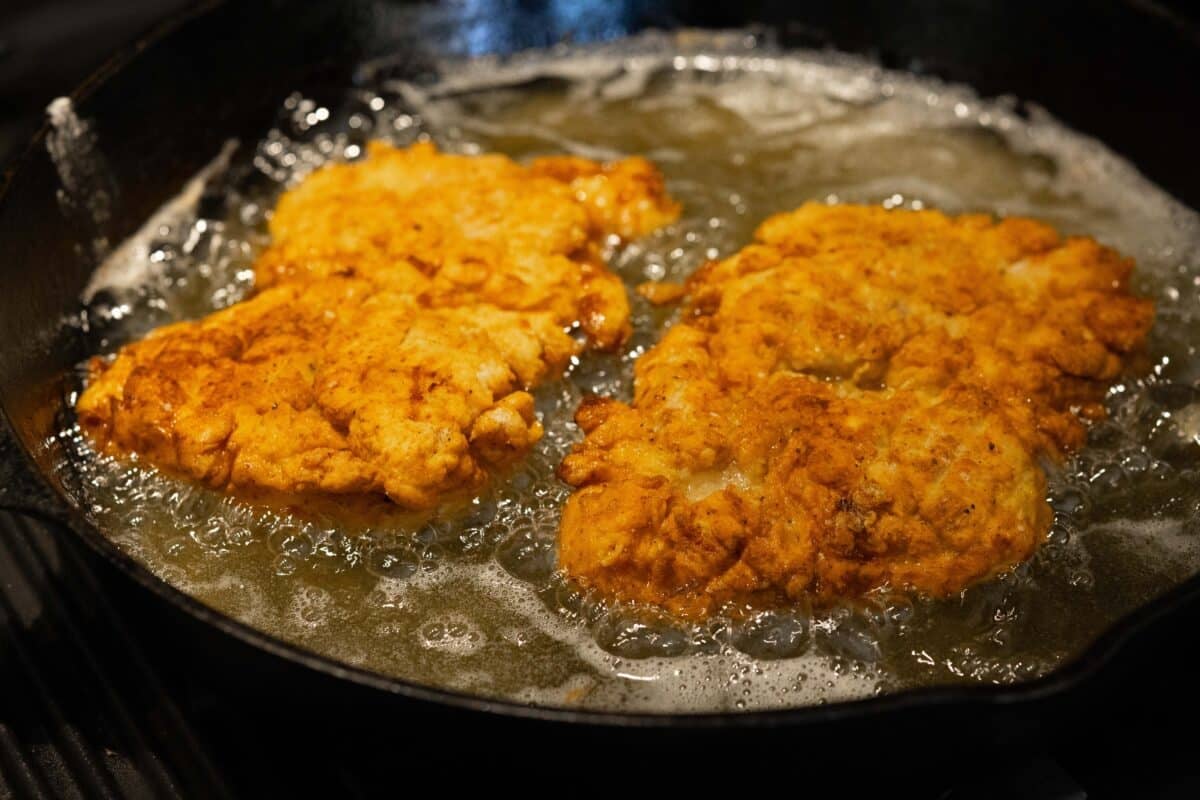 Two breaded chicken thighs frying in oil in a cast-iron pan.