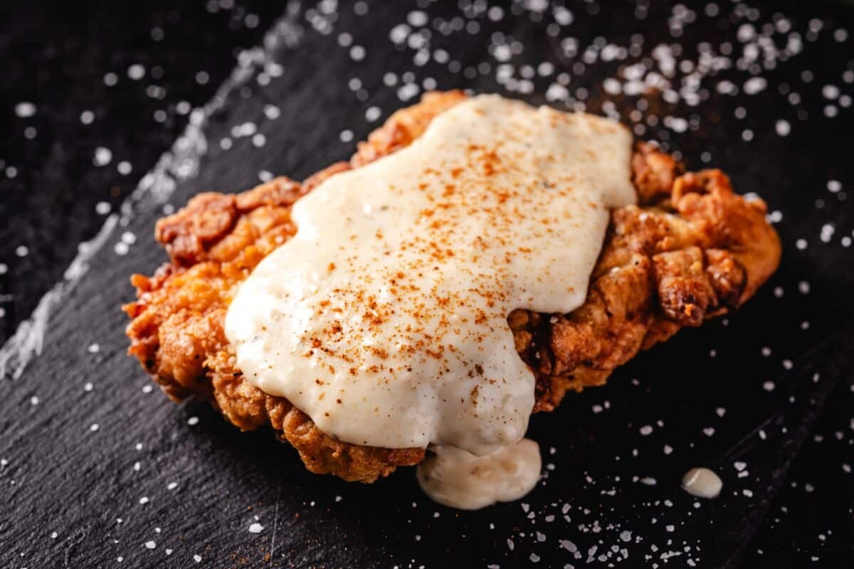 Fried chicken topped with white gravy on a black plate sprinkled with flakes of salt.