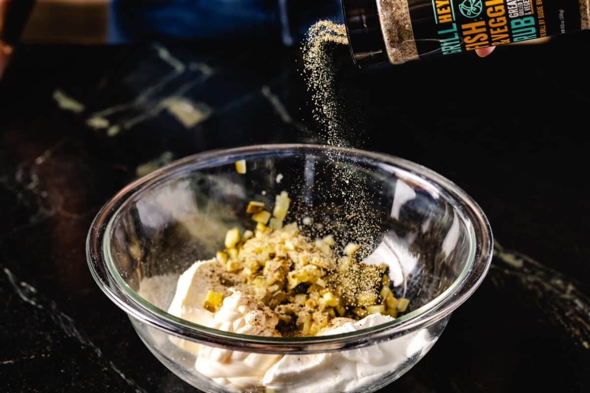 A bottle of rub being sprinkled into a clear bowl filled with mayo and diced pickles.