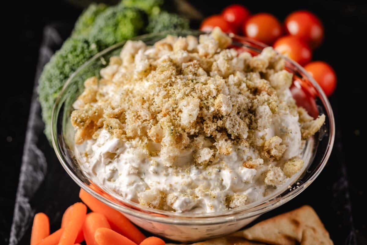 A closeup shot of a bowl of dip next to carrot sticks, broccoli, and tomatoes.