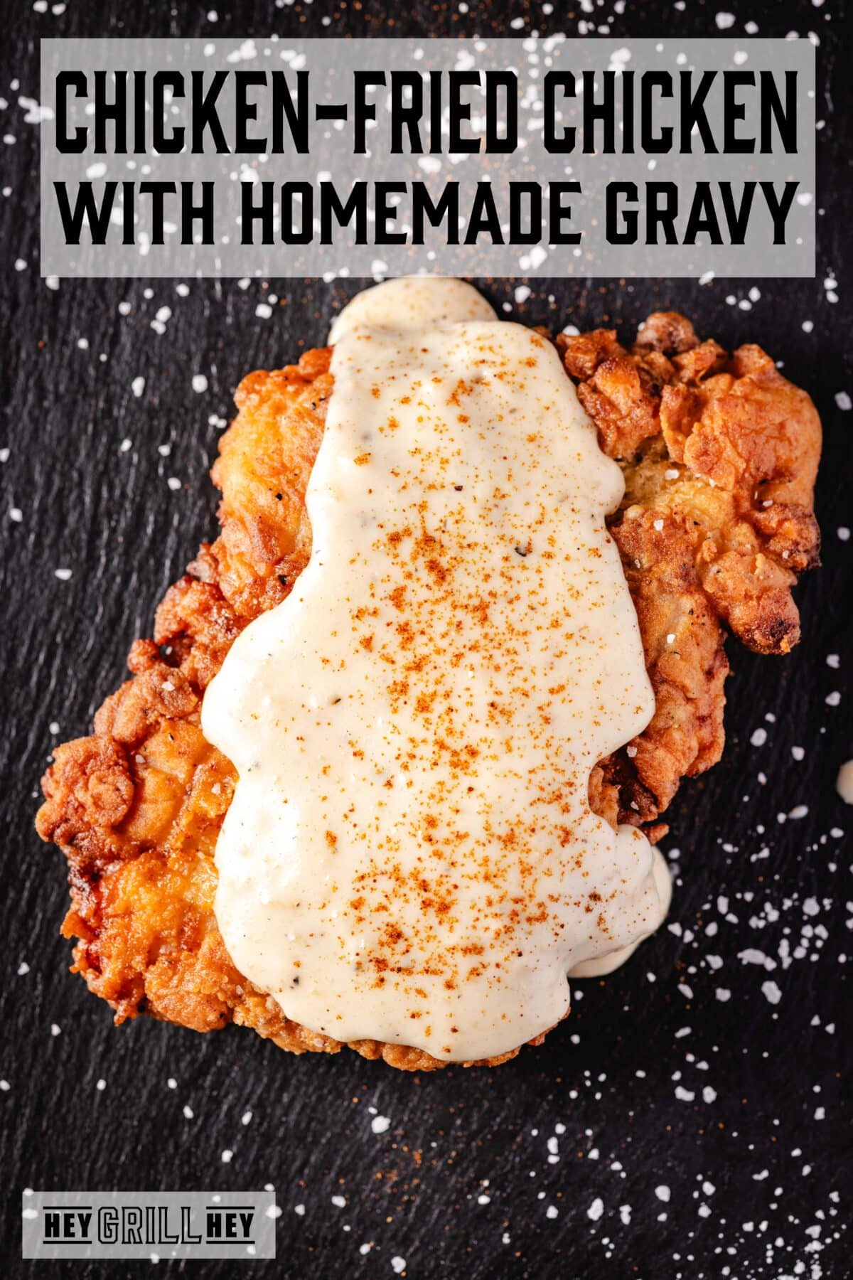 Fried chicken topped with gravy on a black plate sprinkled with flaky salt. The text overlay reads "Chicken Fried Chicken with Homemade Gravy" at the top, and "Hey Grill Hey" at the bottom.