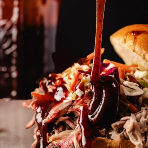 Sauce pouring onto a pulled pork sandwich in front of a jar of the same sauce.