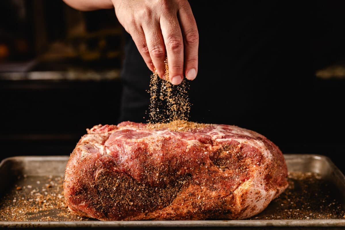A chef's hand sprinkling a dry rub onto a pork shoulder.