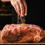 A chef's hand sprinkling a dry rub onto a pork shoulder.