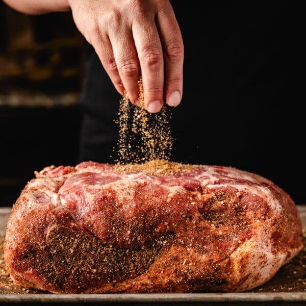 A chef's hand sprinkling a dry rub onto a pork shoulder.