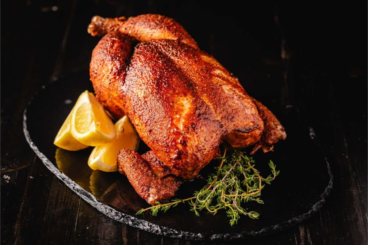 A smoked chicken on a black plate garnished with lemon wedges and herbs.