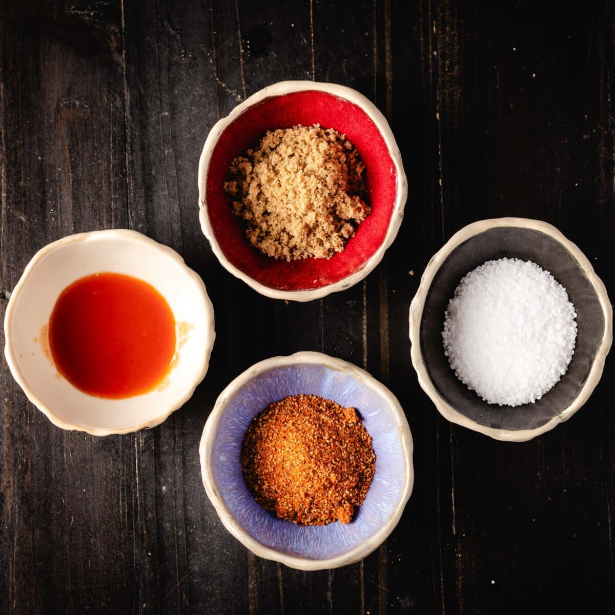 An overhead shot of four bowls containing (clockwise from top) brown sugar, salt, BBQ rub, and hot sauce.