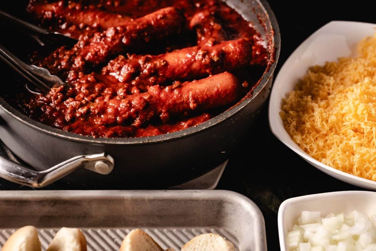 A pot full of chili pepper and hot dogs, a bowl of grated cherdar, a bowl of diced onions and a pan with sandwiches.