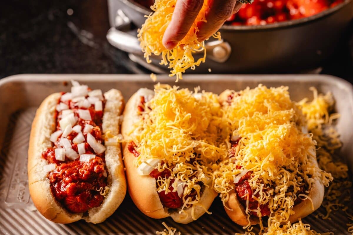 The hand of a chef who sprays the grated cheese on hot dogs on a pan.