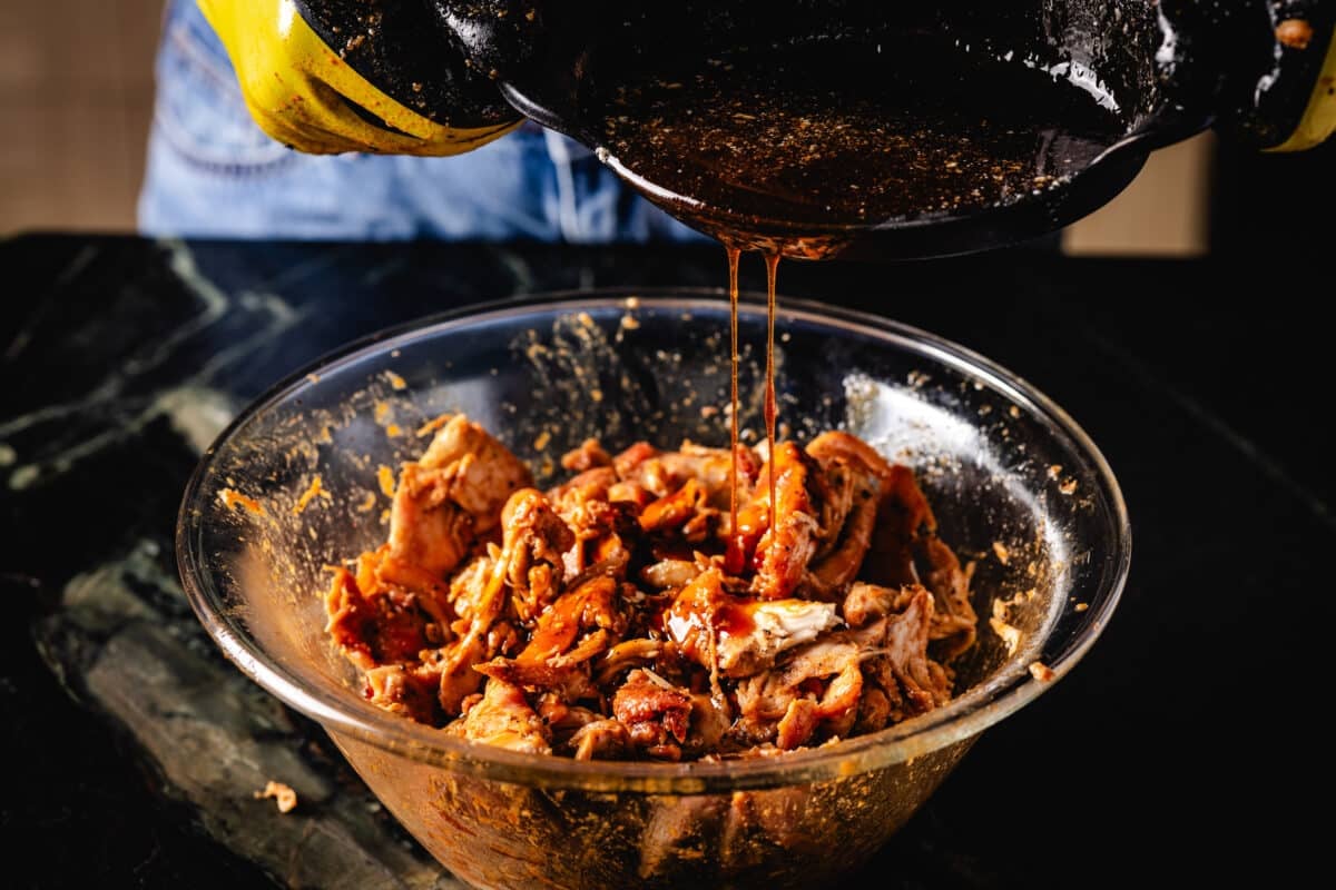 A chef's hands pouring juice from a cast iron skillet into a clear bowl of smoked poultry.