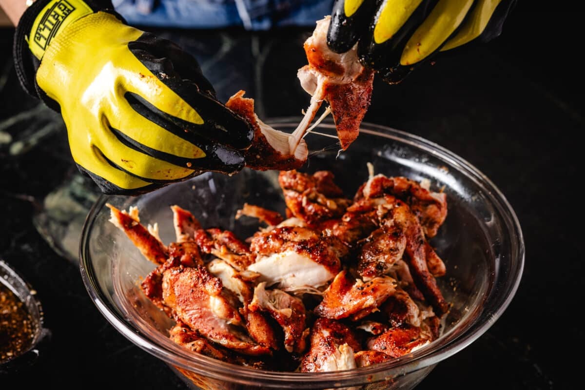 A chef's gloed hands shredding cooked chicken in a glass bowl.