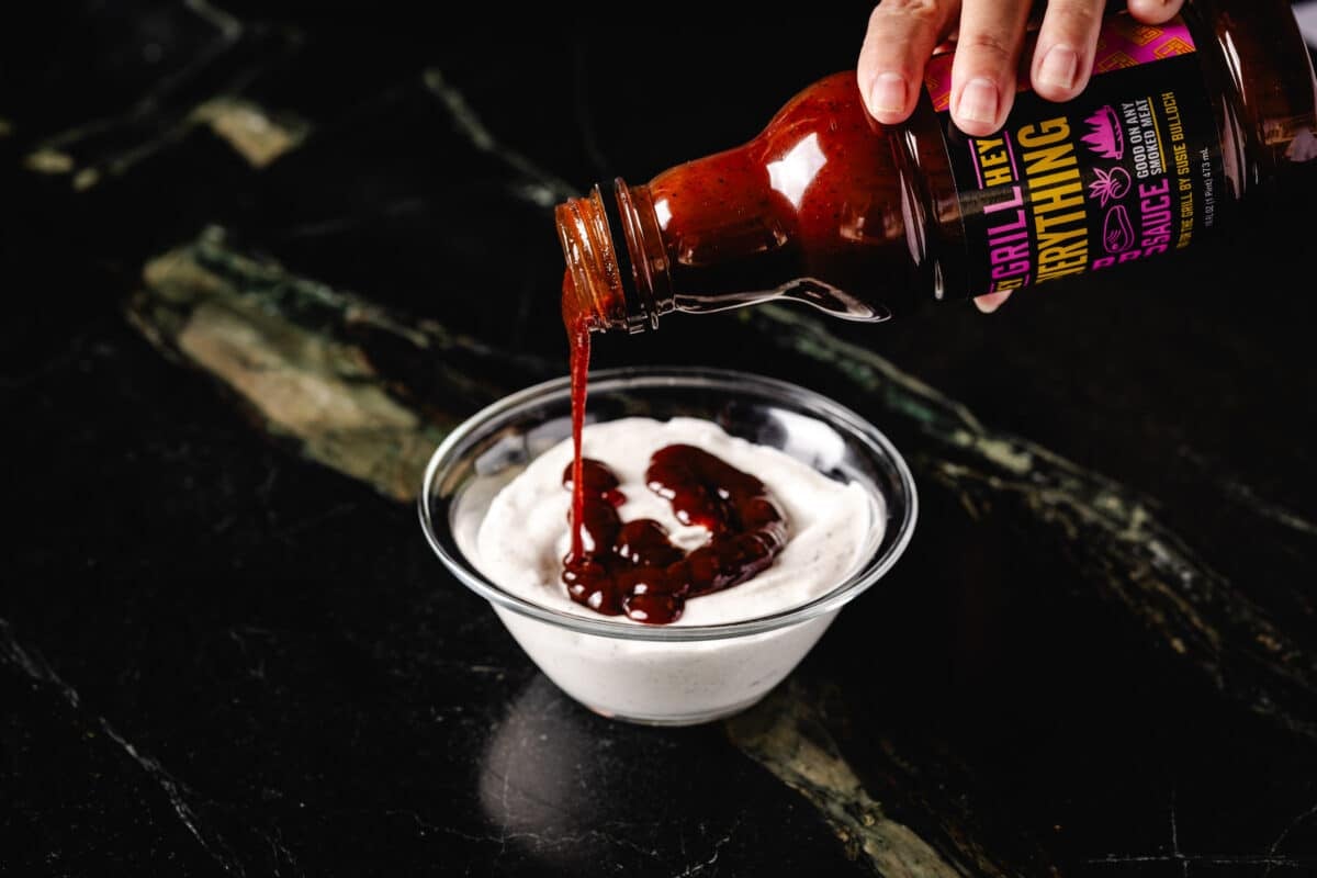 A hand pouring Hey Grill Hey Everything Sauce from a bottle into a bowl of white dressing.