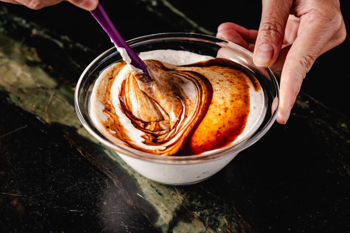 A chef's hand holding a bowl of ranch dressing in place as a rubber spatula stirs in BBQ sauce.