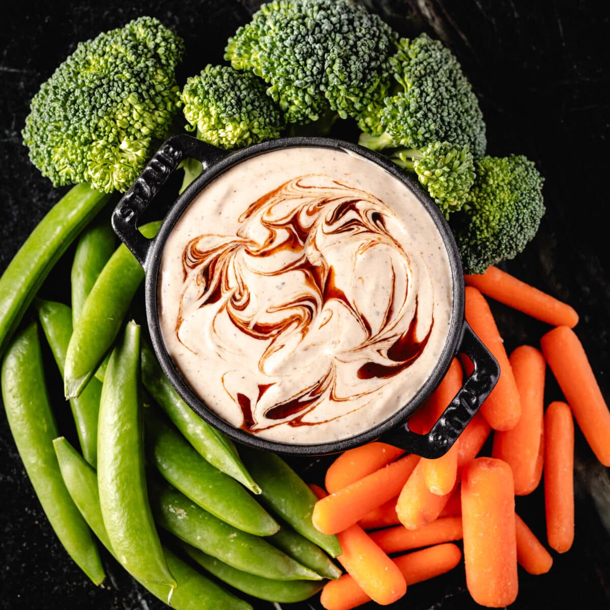 A black bowl filled with ranch dip, surrounded by fresh carrot sticks, broccoli, and snap peas.