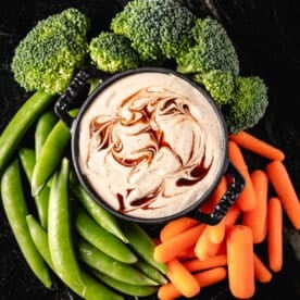 A black bowl filled with ranch dip, surrounded by fresh carrot sticks, broccoli, and snap peas.