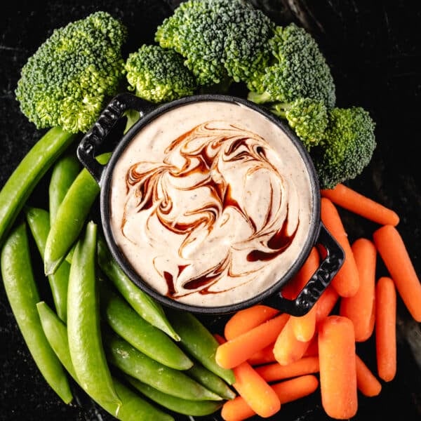 A black bowl filled with ranch dip, surrounded by fresh carrot sticks, broccoli, and snap peas.
