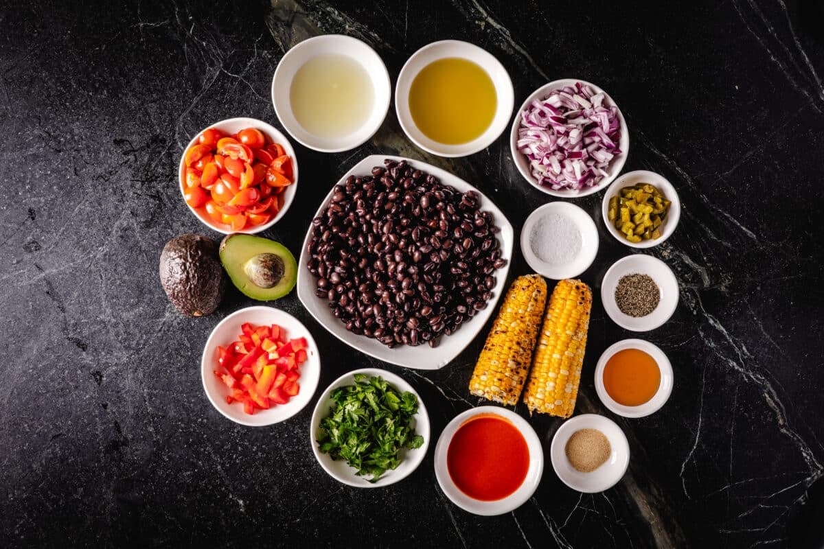 A header of a bowl of beans surrounded by multiple bowls of loose ingredients and ingredients, including corn, onions, tomatoes and condiments.