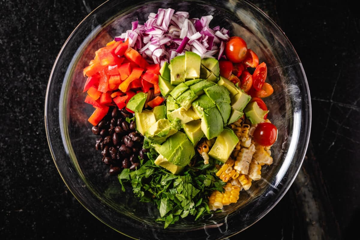 A header of a transparent bowl full of chopped avocado, onion, tomato, coriander, corn, beans and a pepper.
