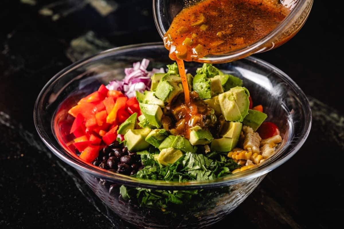 A bowl of brown dressing that pours into a larger bowl full of chopped vegetables.