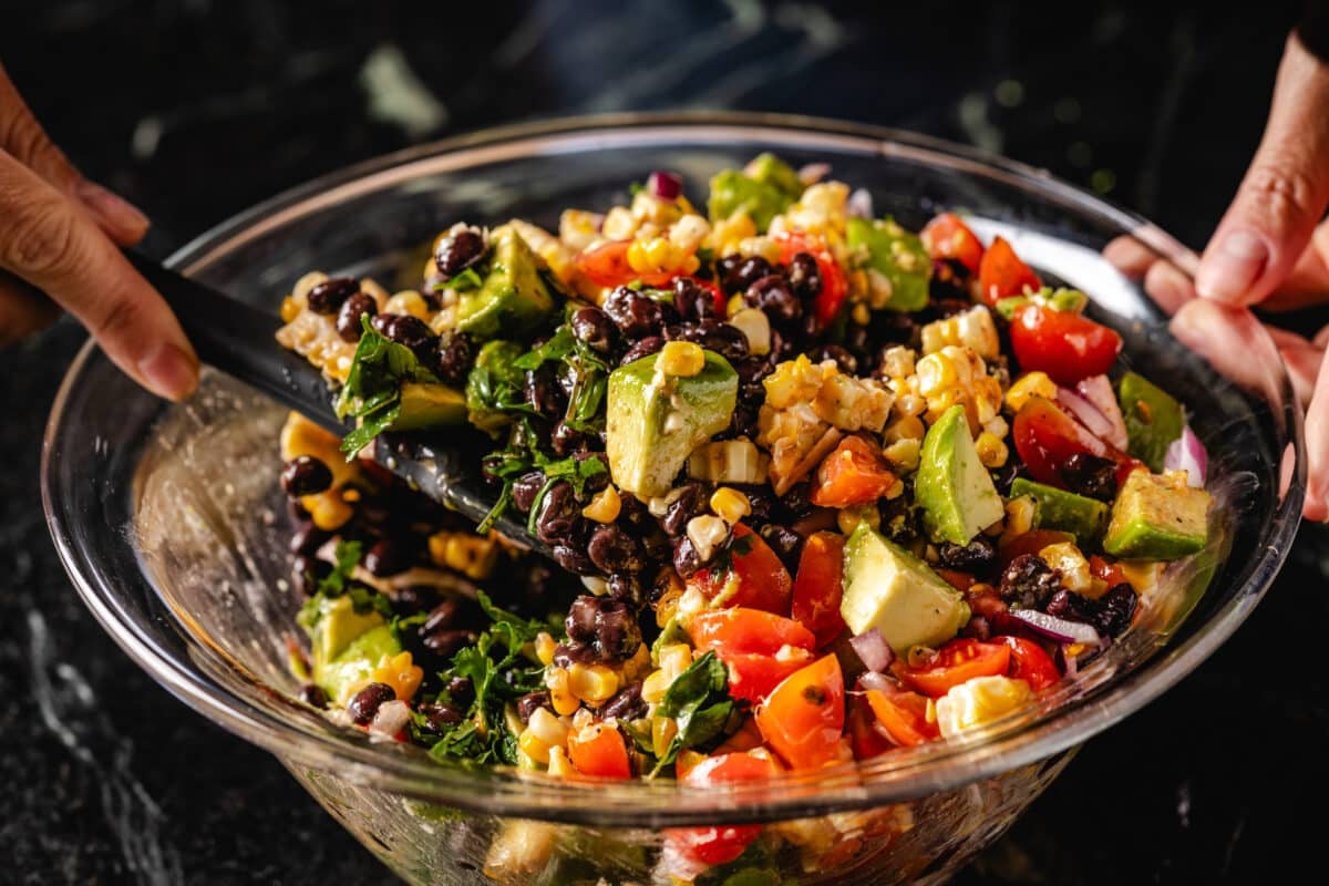 The hands of a chef who use a rubber spatula to mix a vegetable salad in a clear bowl.