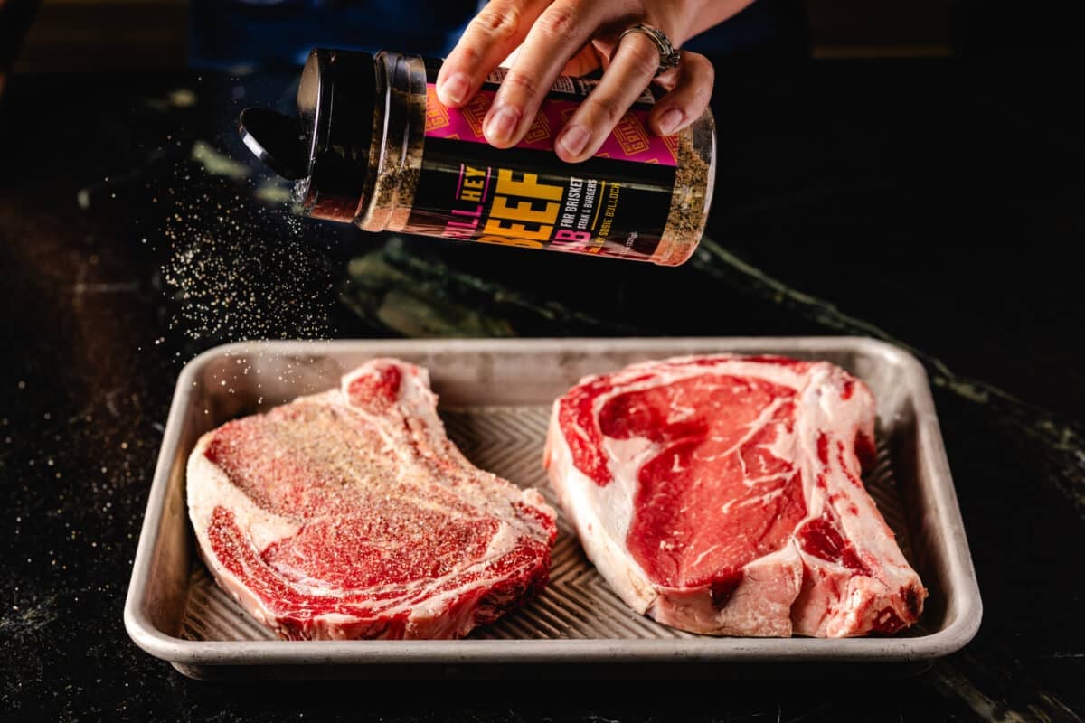 A chef's hand sprinkling Hey Grill Hey Beef Rub onto two raw steaks that are resting on a baking sheet.