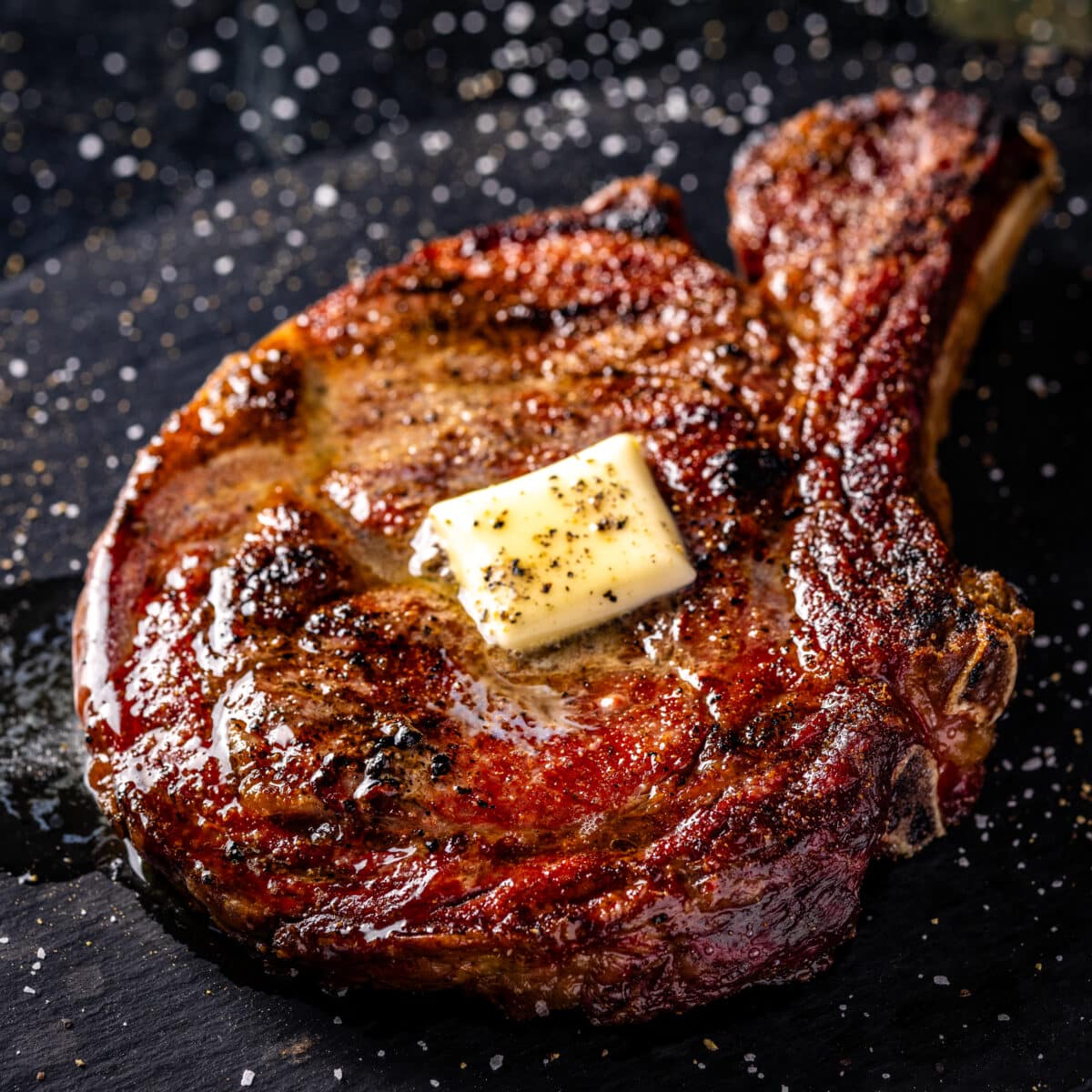 A seared bone-in ribeye steak with a pat of resting butter sitting on a black plate.