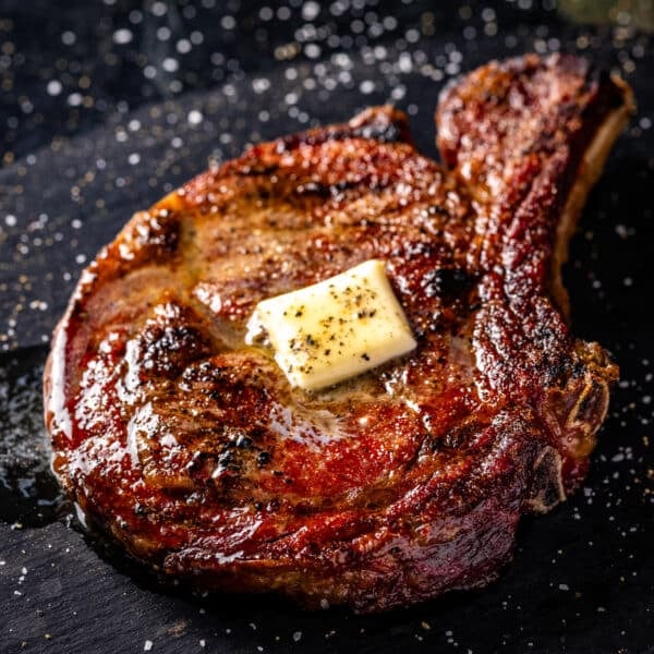 A seared bone-in ribeye steak with a pat of resting butter sitting on a black plate.