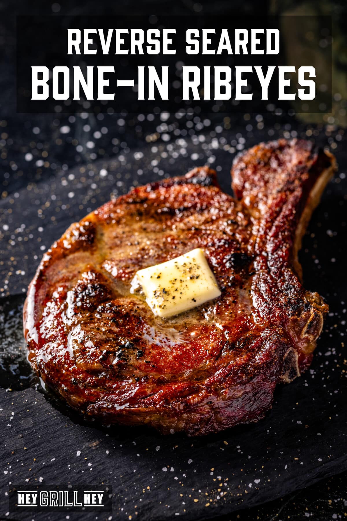 A steak topped with a pat of butter resting on a black plate. The text overlay reads "Reverse Seared Bone-In Ribeyes" at the top, and "Hey Grill Hey" at the bottom.