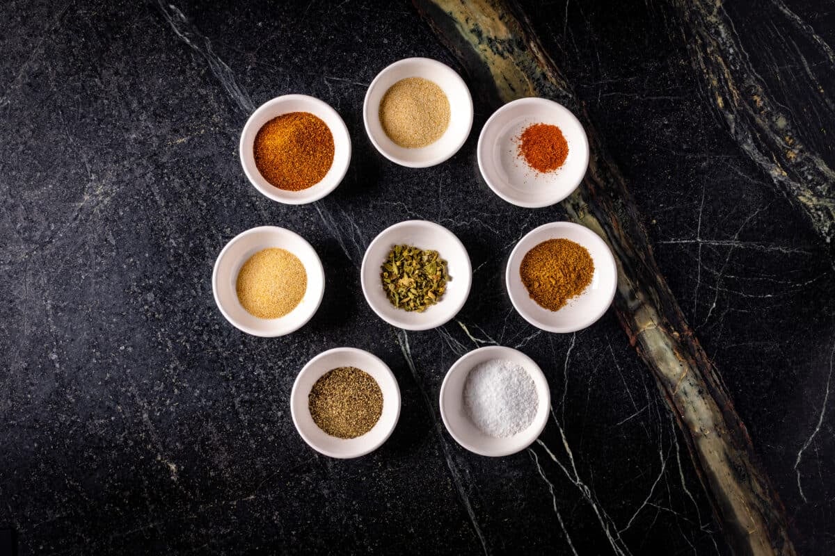 Seven white bowls circled around an eighth bowl, each filled with different spices.