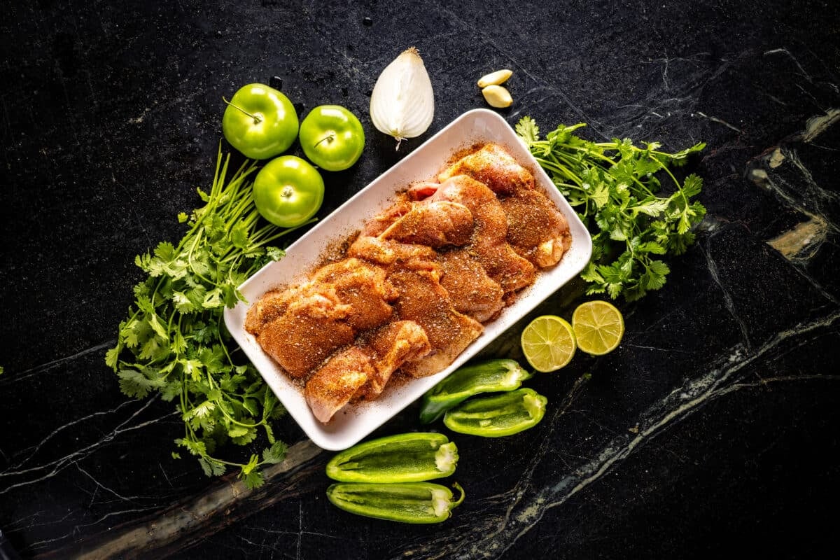 A white platter of seasoned chicken surrounded by lime wedges, tomatillos, cilantro leaves, an onion wedge, and other taco ingredients.