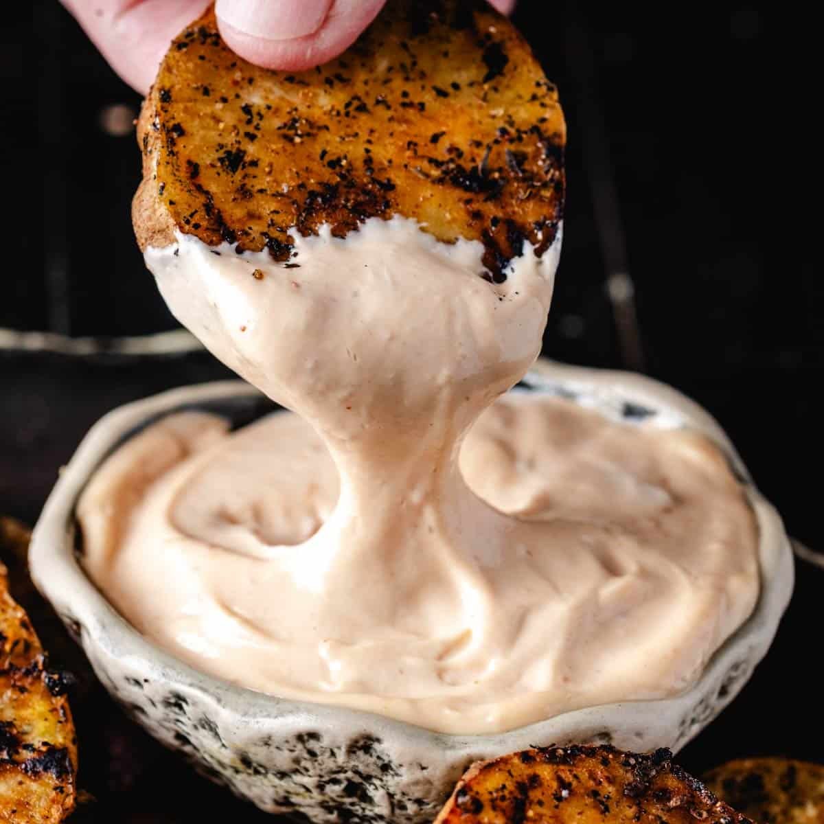 A hand pulling a grilled sweet potato chip into a bowl of dipping sauce.
