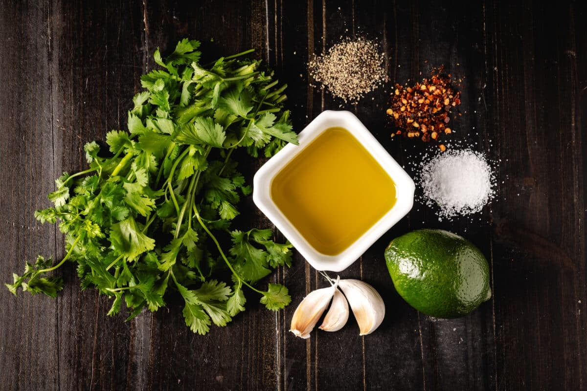 A bunch of fresh cilantro, a bowl of olive oil, bulbs of garlic, a lime, and three piles of seasonings on a dark, wooden surface.
