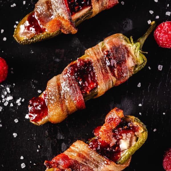 Three bacon wrapped jalapeños on a black platter garnished with flaky salt and fresh berries.