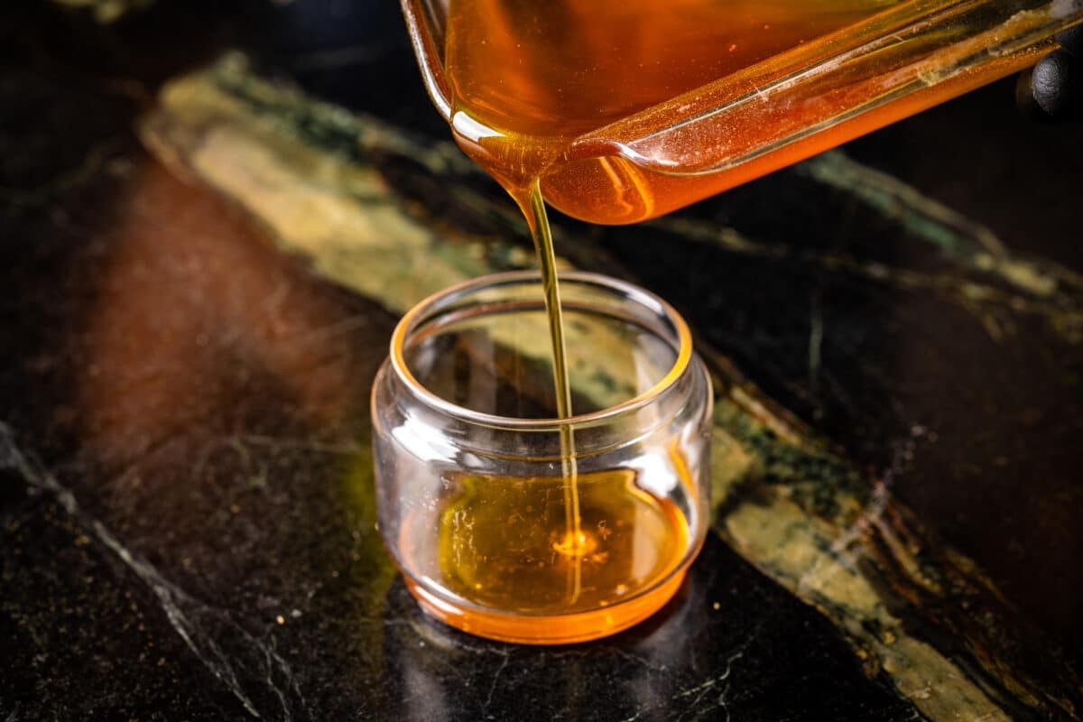 A translucent golden liquid that is poured from a pan into a transparent jar sitting on a marble work surface.