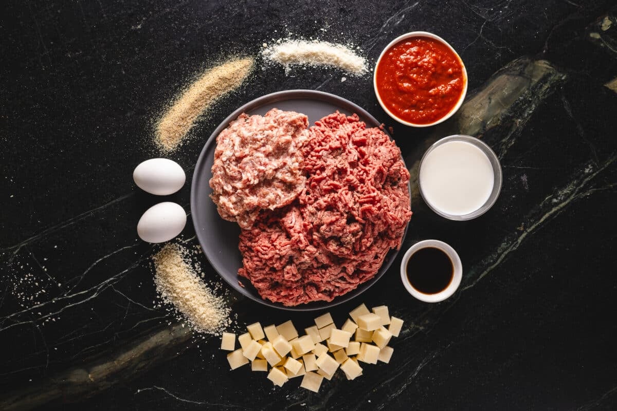 An overhead shot of a plate of ground beef and pork sausage encircled by cheese, eggs, seasonings, and bowls with sauces.