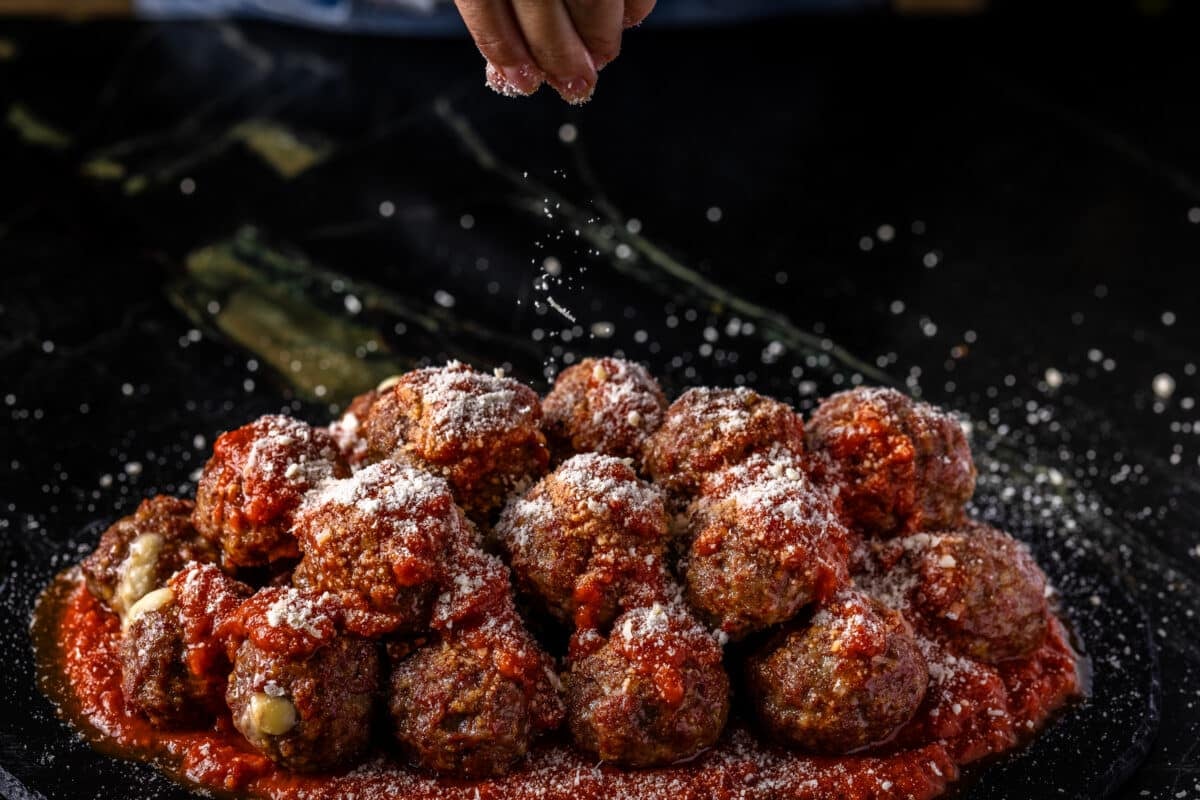 A chef's hand sprinkling grated Parmesan onto a pile of stuffed meatballs.