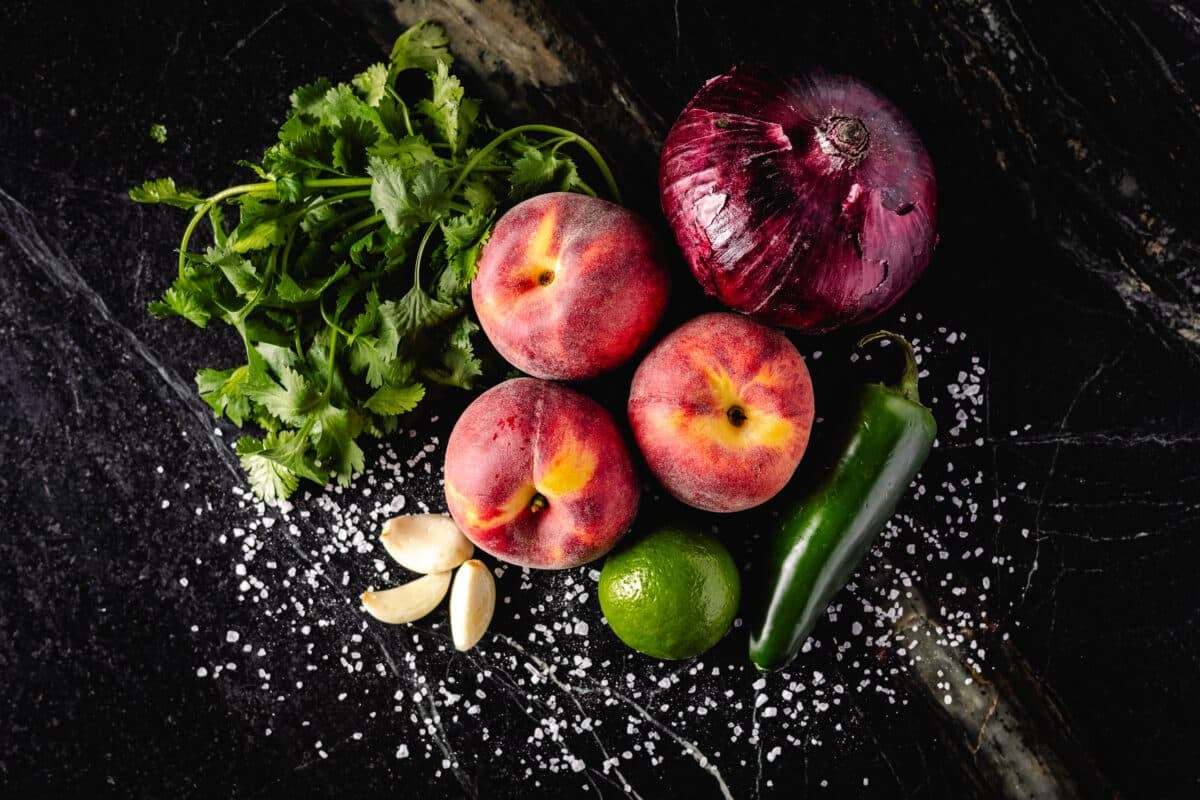 An overhead shot of peaches, cilantro leaves, garlic, a jalapeño, and a lime on a black countertop.