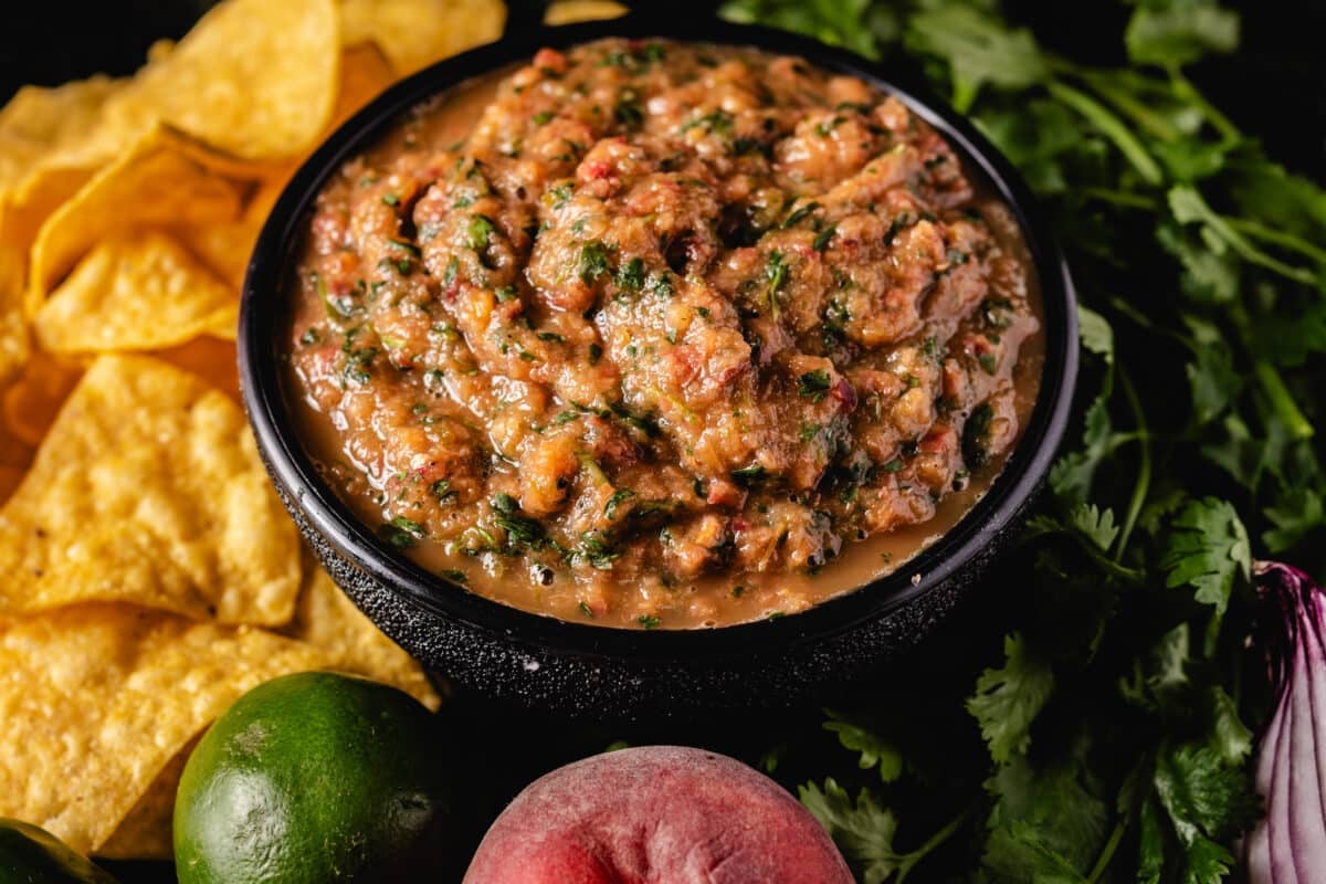 A black bowl filled with salsa next to tortilla chips and raw ingredients.