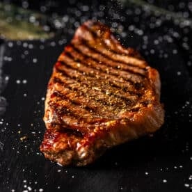 A grilled steak on a black surface being sprinkles with seasoning.