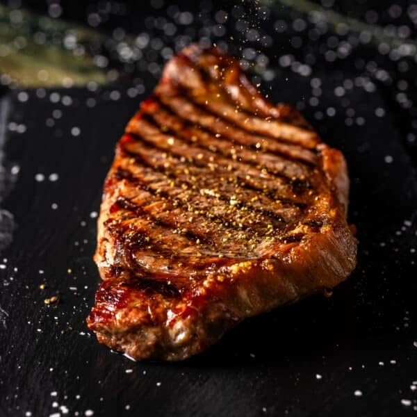 A grilled steak on a black surface being sprinkles with seasoning.